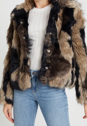 Kvinde iført en brun og sort plettet pelsjakke over en hvid ribbet top og blå højtaljede jeans, stående med afslappede hænder.