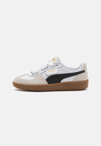Weiße und schwarze Wildleder- und Leder-Sneaker mit einer Gummisohle, ausgestattet mit weißen Schnürsenkeln und gelber Markenkennzeichnung auf der Zunge. Niedrig geschnittenes Design.