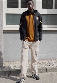 Schwarze Reißverschlussjacke mit gelben Akzenten, orange Polohemd, beiger Cargo-Hose und schwarze Adidas Sneaker mit weißen Streifen.