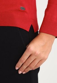 Pull en tricot rouge avec des poignets et un ourlet côtelés, mettant en avant un petit logo en métal. Le bas présente un léger design fendu.