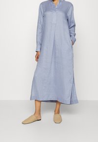 Kobieta w długiej, jasnoniebieskiej lnianej sukience z długimi rękawami i beżowych, plecionych butach wsuwanych, stojąca na jednolitym tle.
