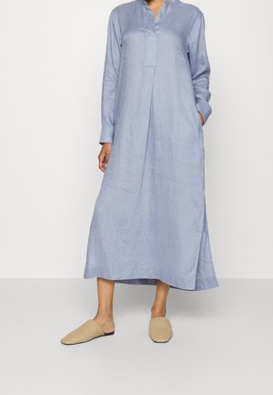 Kvinde iført en lang lyseblå linnedkjole med lange ærmer og beige vævede slips-on sko, stående mod en ensfarvet baggrund.