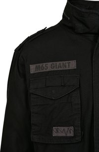 Zwarte M65-jack met een hoge kraag, voorzien van twee voorzakken en een patch met het label "M65 GIANT." De stof heeft een gestructureerde afwerking.