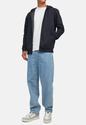 Donkerblauwe ritshoodie met twee voorzakken, gecombineerd met lichtblauwe baggy jeans en grijs-witte sneakers. Zachte katoenen stof.
