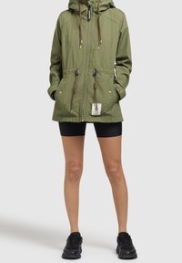 Persona che indossa una giacca con cappuccio verde oliva con coulisse, pantaloni corti da ciclismo neri e sneaker nere, in piedi contro uno sfondo grigio.