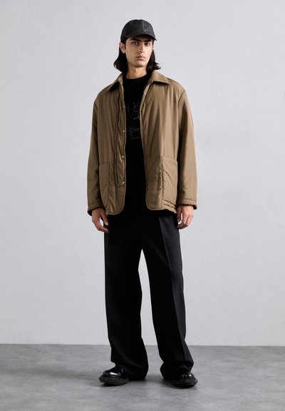 Chaqueta acolchada marrón con cuello de punta, dos bolsillos frontales, llevada sobre una camiseta gráfica negra y combinada con pantalones negros holgados y zapatos negros.