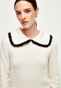 Pull créme avec un grand col noir à volants. La texture semble douce, avec des manches longues et une coupe ajustée.