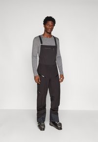 Juodos sniego kelnės su reguliuojamais užtrauktukais, kišenėmis ir pritaikytu dizainu. Suderinta su pilku ilgomis rankovėmis marškinėliais ir juodais slidinėjimo batais.