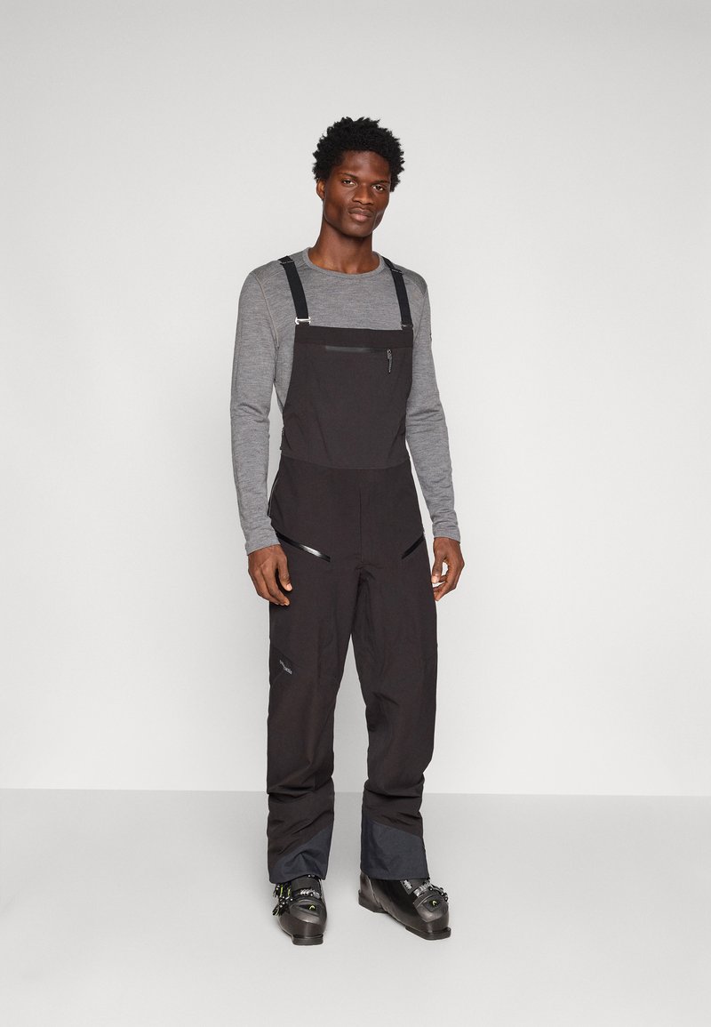 Juodos sniego kelnės su reguliuojamais užtrauktukais, kišenėmis ir pritaikytu dizainu. Suderinta su pilku ilgomis rankovėmis marškinėliais ir juodais slidinėjimo batais.