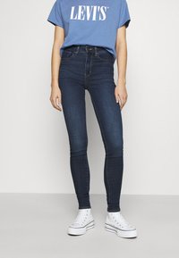 Jeans oscuros de corte slim, combinados con una camiseta azul de Levi's metida por dentro y zapatillas altas blancas, de pie contra un fondo liso.