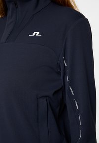Pullover in pile blu navy con mezza cerniera e superficie testurizzata. Presenta un logo bianco sul petto e dettagli con marchio lungo le maniche.