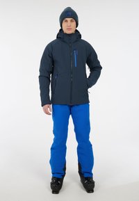 Donkerblauwe skijas met een rits aan de voorkant en een capuchon, met een borstzak met blauwe accenten. Gecombineerd met heldere blauwe skibroeken en zwarte skischoenen.