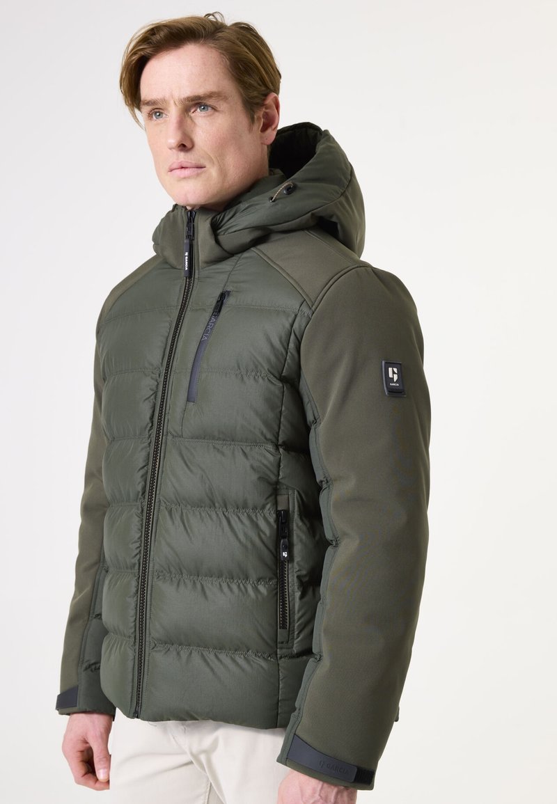 Olivgrüne Steppjacke mit abnehmbarem Kapuzen, Reißverschlusstaschen, gerippten Manschetten und strukturiertem Finish; die Polsterung ist im gesteppten Design sichtbar.