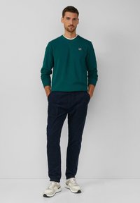 Sudadera color verde azulado con escote redondo y logo, combinada con pantalones azul marino oscuro. Zapatillas deportivas en tonos beige y blanco con acentos en verde completan el look.