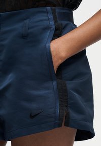Κοντινό σε χέρι ατόμου στην τσέπη σορτς Nike σε ναυτικό μπλε με μαύρη δαντέλα και διακοσμητικές λεπτομέρειες.