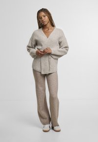 Světle béžový zavinovací svetr vyrobený z měkkého materiálu, s výstřihem do V, širokými rukávy a stahováním v pase, doplněný o odpovídající kalhoty.