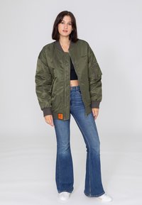 Veste bomber vert olive avec fermeture éclair à l'avant, poignets côtelés et étiquette orange. Assortie à un jean bleu évasé et des chaussures blanches.