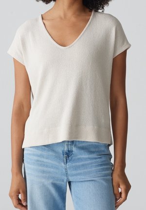 Vrouw draagt een lichtbeige, kort mouw gebreide top met V-hals, gecombineerd met lichtblauwe spijkerbroek tegen een effen achtergrond.