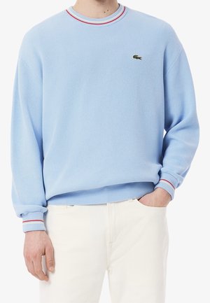 Mężczyzna w jasnoniebieskim swetrze z okrągłym dekoltem z czerwono-białymi paskami na kołnierzu i mankietach oraz w białych spodniach, z ręką w kieszeni