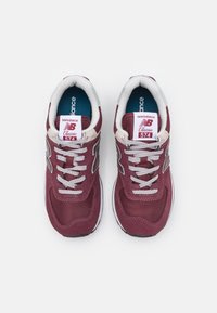 Sneakers New Balance 574 color bordeaux con materiali in mesh e suede, lacci grigi, dettagli bianchi, colletto imbottito e etichetta con marchio sulla linguetta.