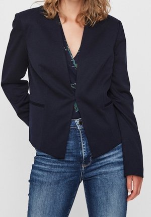 Kvinde iført en mørk marineblazer over en blomsterprintet top kombineret med blå denimjeans, poserer med én hånd bag ryggen.