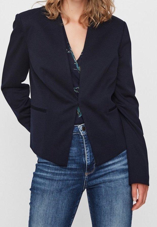 Femme portant un blazer bleu marine foncé sur un haut fleuri associé à un jean en denim bleu, posant avec une main dans le dos.