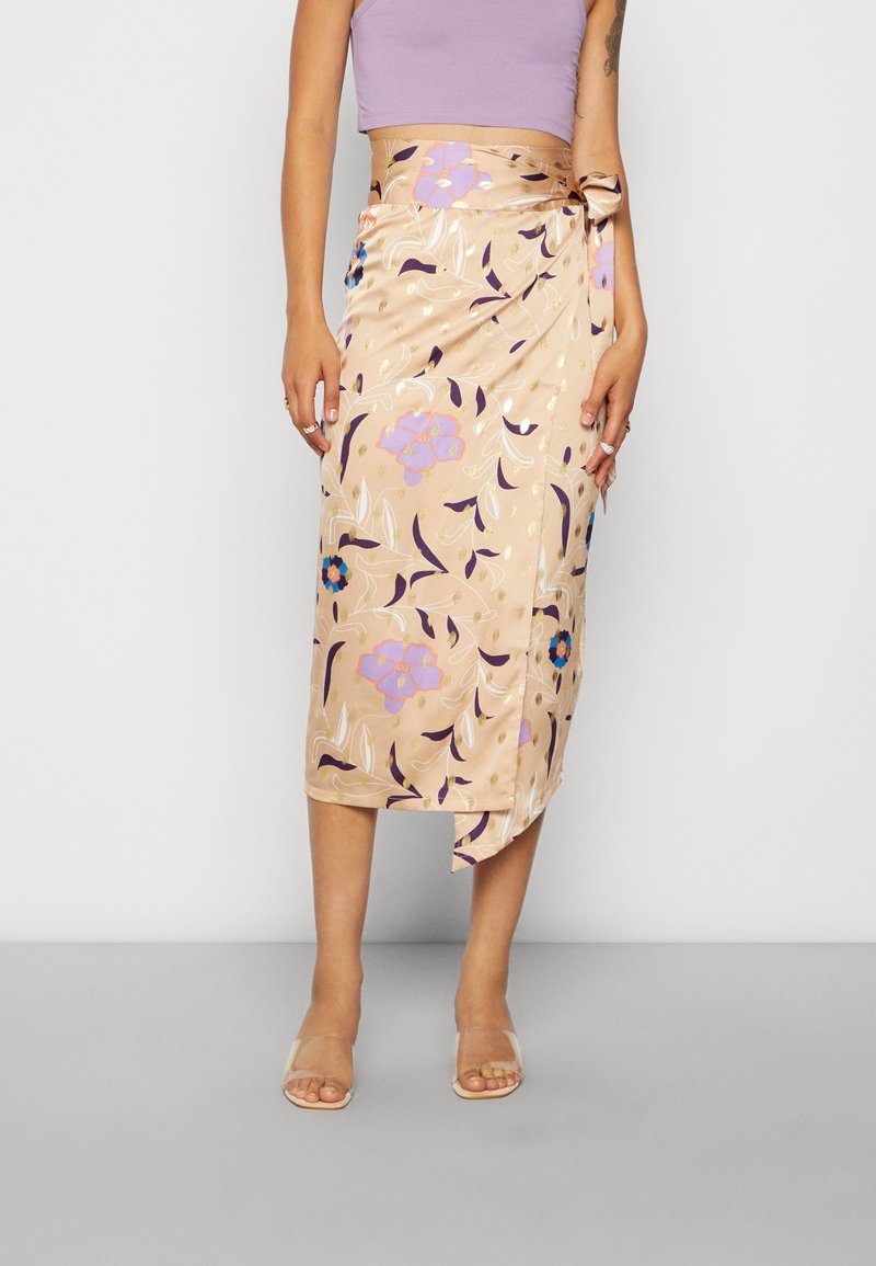 Femme portant une jupe midi portefeuille en satin beige avec un imprimé floral violet et bleu, associée à un top court lavande sans manches et des sandales transparentes.