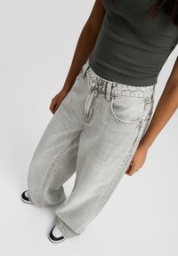 Femme portant un jean ample délavé clair, un débardeur noir et des baskets noir et blanc, debout devant un fond clair uni.
