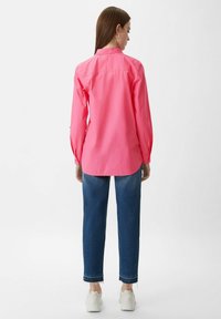 Roze langwerpige blouse met een comfortabele pasvorm, knopenmanchetten en een gebogen zoom, gecombineerd met blauwe jeans en witte sneakers.