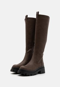 Bottes brunes montant jusqu'aux genoux en matériau synthétique texturé, dotées d'une fermeture éclair latérale et d'une semelle plateforme noire épaisse avec des crampons profonds.