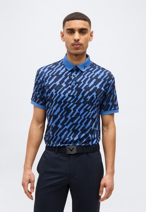 Mlajši moški stoji, oblečen v modro polo majico s kratkimi rokavi in vzorcem ter temne hlače s črnim pasom.