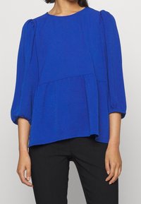 Blusa blu, con texture, dalla scollatura rotonda, maniche a sbuffo e un design a strati, abbinata a pantaloni neri.