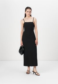 Robe maxi noire sans manches avec un décolleté carré, texture lisse et silhouette ajustée. Portée avec des sandales noires à lanières et un petit sac.