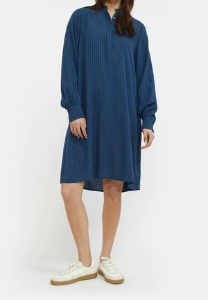 Femme portant une robe bleu marine ample à manches longues et longueur genou, avec des rayures verticales subtiles, ainsi que des baskets blanches.