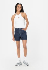 Hvit halterneck-topp med grafisk logo, kombinert med blå denimshorts. Modellen har på seg grå sneakers og hvite ankelsokker. Enkelt design, avslappet utseende.