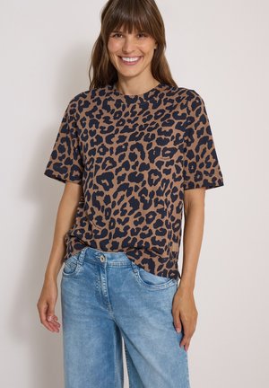 Femme souriante portant un t-shirt à manches courtes imprimé léopard marron et noir et un jean bleu clair sur un fond uni clair.