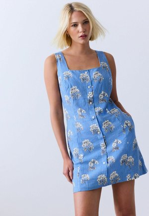 Jonge vrouw met blond haar die een blauwe mouwloze jurk draagt met witte bloemmotieven en knopen aan de voorkant, staand voor een effen achtergrond.
