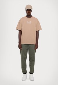 Barna, oversized póló fehér logóval, zöld, tapered fazonú nadrág és fehér tornacipő. A sapka illeszkedik a póló színéhez, így komplett casual szettet alkot.