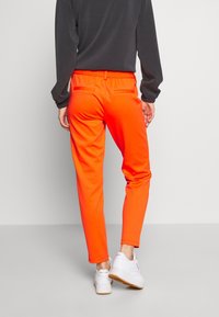Felle oranje trainingsbroek met een elastische tailleband, taps toelopende pasvorm en twee achterzakken, gecombineerd met witte sneakers op een grijze achtergrond.