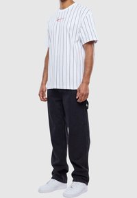 T-shirt bianco a maniche corte con strisce verticali nere e logo rosso, abbinato a pantaloni neri e sneakers bianche. Design semplice e casual.