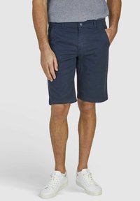Marineblaue Baumwollshorts mit geradem Schnitt, ausgestattet mit Seitentaschen und einem Knopfverschluss. Kombiniert mit weißen Turnschuhen.