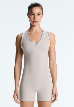 Vrouw staat met het gezicht naar voren en draagt een beige, mouwloze, aansluitende romper met lichtblauwe biezen bij de halslijn en armsgaten.