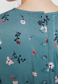 Grön tyg med blommönster och små knappar på baksidan. Innehåller delikata blommor i rosa och vitt mot en blågrön bakgrund.