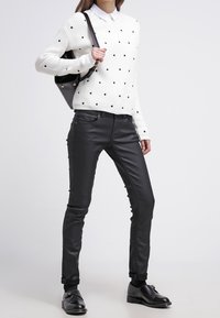 Vit stickad tröja med svarta prickar, ihop med svarta smalpassande byxor i konstläder och svarta skor. Modellen håller en svart väska.