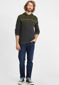 Gebreide hoodie in donker groen en grijs met een gestructureerd patroon; heeft een capuchon met trekkoord en een slanke pasvorm. Gecombineerd met donkerblauwe spijkerbroek en canvas sneakers.