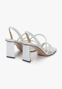 Witte sandalen met hakken en vierkante blokhakken, gekruiste banden en een gladde leren afwerking. De zool heeft een lichte kromming, met zwarte accenten.