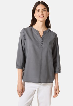 Graues Langarmshirt mit V-Ausschnitt und Knopfleiste mit drei Knöpfen; glattes Material mit lockerer Passform; kombiniert mit weißen Hosen.