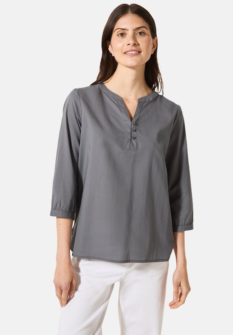 Camicia grigia a maniche lunghe con scollo a V e patta con tre bottoni; tessuto morbido con una vestibilità comoda; abbinata a pantaloni bianchi.