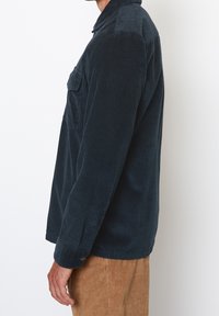 Granatowa koszula sztruksowa z klasycznym kołnierzem, kieszenią z przodu i mankietami na guziki. Charakteryzuje się pionowym prążkowaniem i luźnym fasonem.