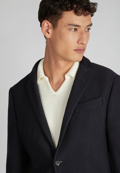 Granatowa marynarka o fakturze z klapami w serek, zapinana na pojedynczy guzik, z kieszonką na piersi, noszona na jasnym, kremowym swetrze z dekoltem w serek.